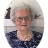 Portrait einer &auml;lteren Frau mit grauem Haar, Brille und Perlenkette vor einer hellen, schlicht dekorierten Wand.