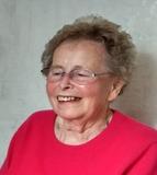 Lächelnde ältere Frau mit Brille in pinkem Pullover vor einer hellen, strukturierten Wand.