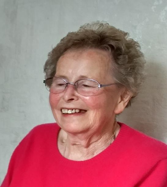 Lächelnde ältere Frau mit Brille in pinkem Pullover vor einer hellen, strukturierten Wand.