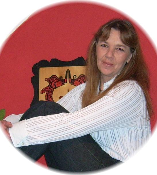 Frau mit langem Haar in gestreiftem Hemd sitzt mit angezogenen Beinen vor roter Wand und einem hölzernen Kunstwerk.