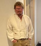 Mann mit Brille trägt eine weiße Bluse und geschnürte beige Hosen in einem hellen Raum mit Holzdetails.