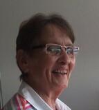 Lächelnde ältere Frau mit Brille und gestreiftem Oberteil, sitzt vor einer hellen Wand.