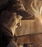 Nahaufnahme eines älteren Mannes mit Bart, der nachdenklich an einem Tisch sitzt, beleuchtet von flackerndem Licht.