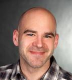 Porträt eines Mannes mit Glatze, in einem karierten Hemd, lächelt vor einem verschwommenen, grauen Hintergrund.