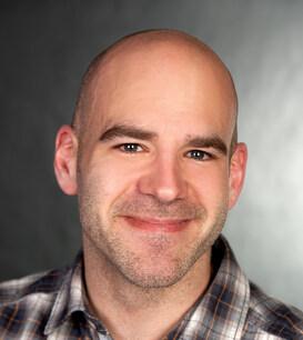 Porträt eines Mannes mit Glatze, in einem karierten Hemd, lächelt vor einem verschwommenen, grauen Hintergrund.