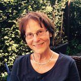 Ältere Frau mit Brille und kurzem Haar, lächelt im Freien vor einem grünen Hintergrund aus Pflanzen.