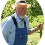 Älterer Mann mit Brille und Strohhut steht smilend im Freien, trägt blaues Hemd und Latzhose, hält einen Stock.