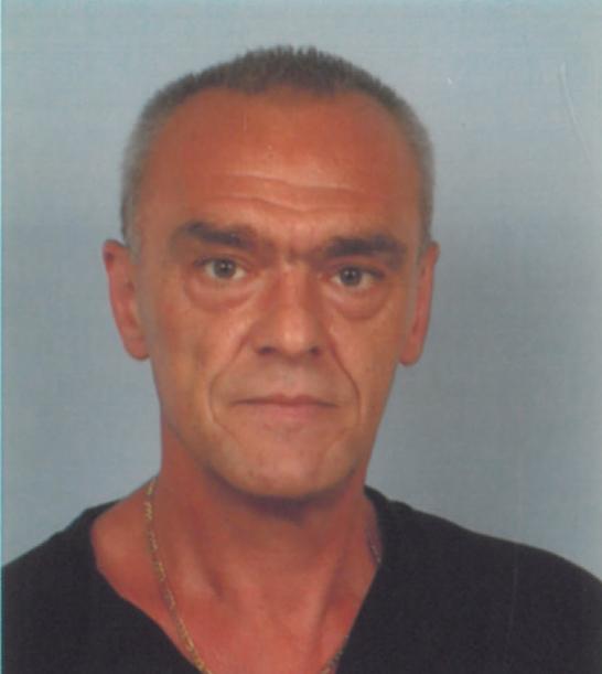 Porträt eines Mannes mit kurzem grauem Haar, goldener Kette und schwarzem Hemd, vor einem hellblauen Hintergrund.