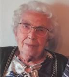 Ältere Frau mit Brille und lockigem, grauem Haar, in einem gemusterten Oberteil, lächelt leicht in die Kamera.