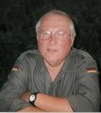 Älterer Mann mit Brille in grauer Uniform, lächelt und hat die Arme verschränkt, Hintergrund unscharf.