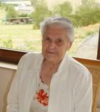 Ältere Frau mit grauen Haaren sitzt an einem Tisch, im Hintergrund eine grüne Landschaft und Blumen.