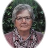 Porträt einer älteren Frau mit Brille und grauen Haaren, umgeben von grüner Vegetation. Sie trägt einen lila Schal.