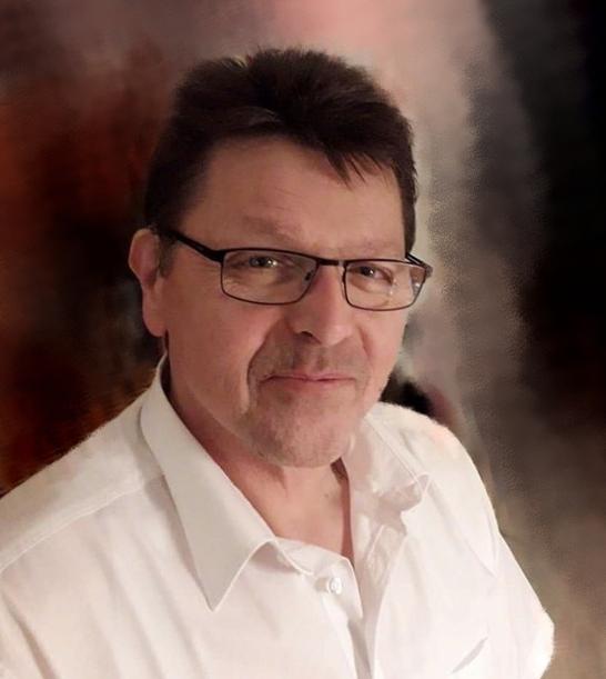 Mann mit Brille und kurzen, dunklen Haaren, trägt ein weißes Hemd vor einem unscharfen, dunklen Hintergrund.