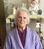 Ältere Frau mit grauen Haaren in lila Strickjacke, lächelt vor dekorativem Hintergrund mit Figuren und Pflanzen.