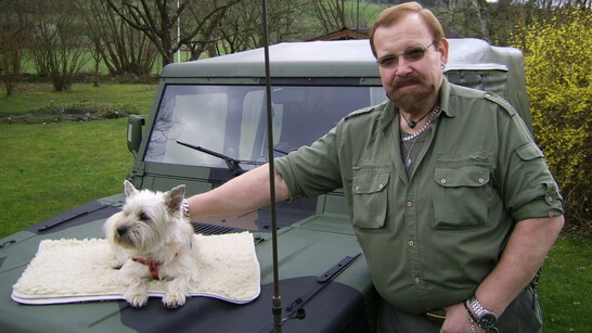 Mann im grünen Outfit steht neben einem Geländewagen, während ein kleiner Hund auf der Motorhaube liegt.