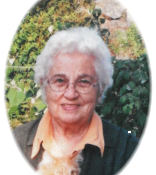 Ältere Frau mit grauem, lockigem Haar lächelt vor einer grünen Landschaft und hält eine orangefarbene Katze im Arm.