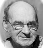 Porträt eines älteren Mannes mit Glatze und Brille, der ernst in die Kamera schaut, vor einem unscharfen Hintergrund.