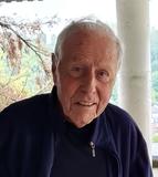 Älterer Mann mit grauen Haaren und freundlichem Lächeln steht vor einem Balkon mit grünem Dach und Bäumen im Hintergrund.