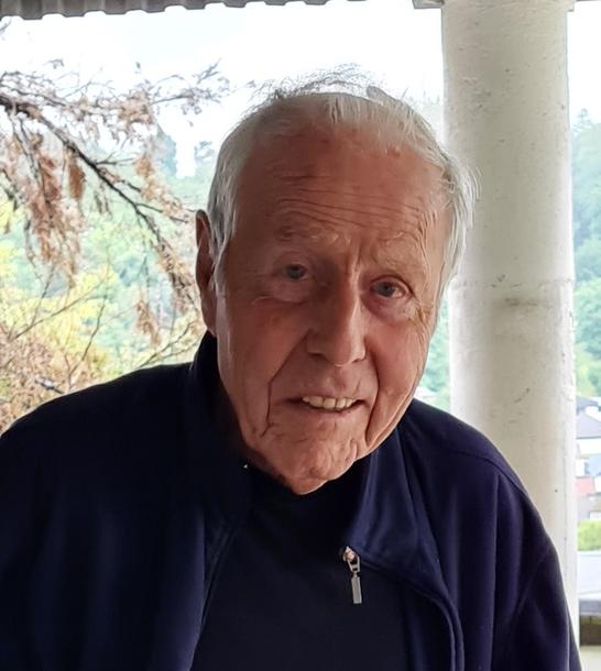 Älterer Mann mit grauen Haaren und freundlichem Lächeln steht vor einem Balkon mit grünem Dach und Bäumen im Hintergrund.