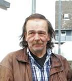 Nahaufnahme eines Mannes mit langen Haaren und einem Bart, der in einer braunen Lederjacke vor einem Gebäude steht.