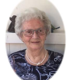 Portrait einer älteren Frau mit grauem Haar, Brille und Perlenkette vor einer hellen, schlicht dekorierten Wand.