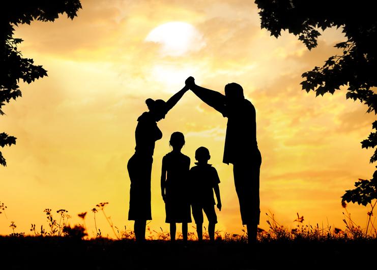 Silhouette einer vierköpfigen Familie, die bei Sonnenuntergang im Freien steht. Die Eltern bilden mit ihren erhobenen Händen ein Dach über ihren zwei Kindern. Bäume rahmen die Szene ein.