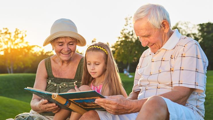 Großeltern und Enkelin sitzen im Freien und lesen gemeinsam in einem Buch bei Sonnenuntergang.