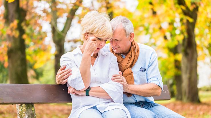 Ein älteres Paar sitzt auf einer Bank im Herbst, mit tröstender Körpersprache und ernsten Gesichtsausdrücken.