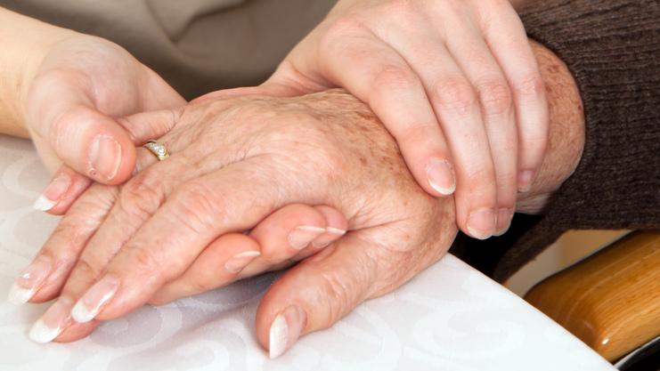 Zwei Hände, eine ältere und eine jüngere, liegen auf einem Tisch, wobei die jüngere Hand die ältere sanft umschließt.